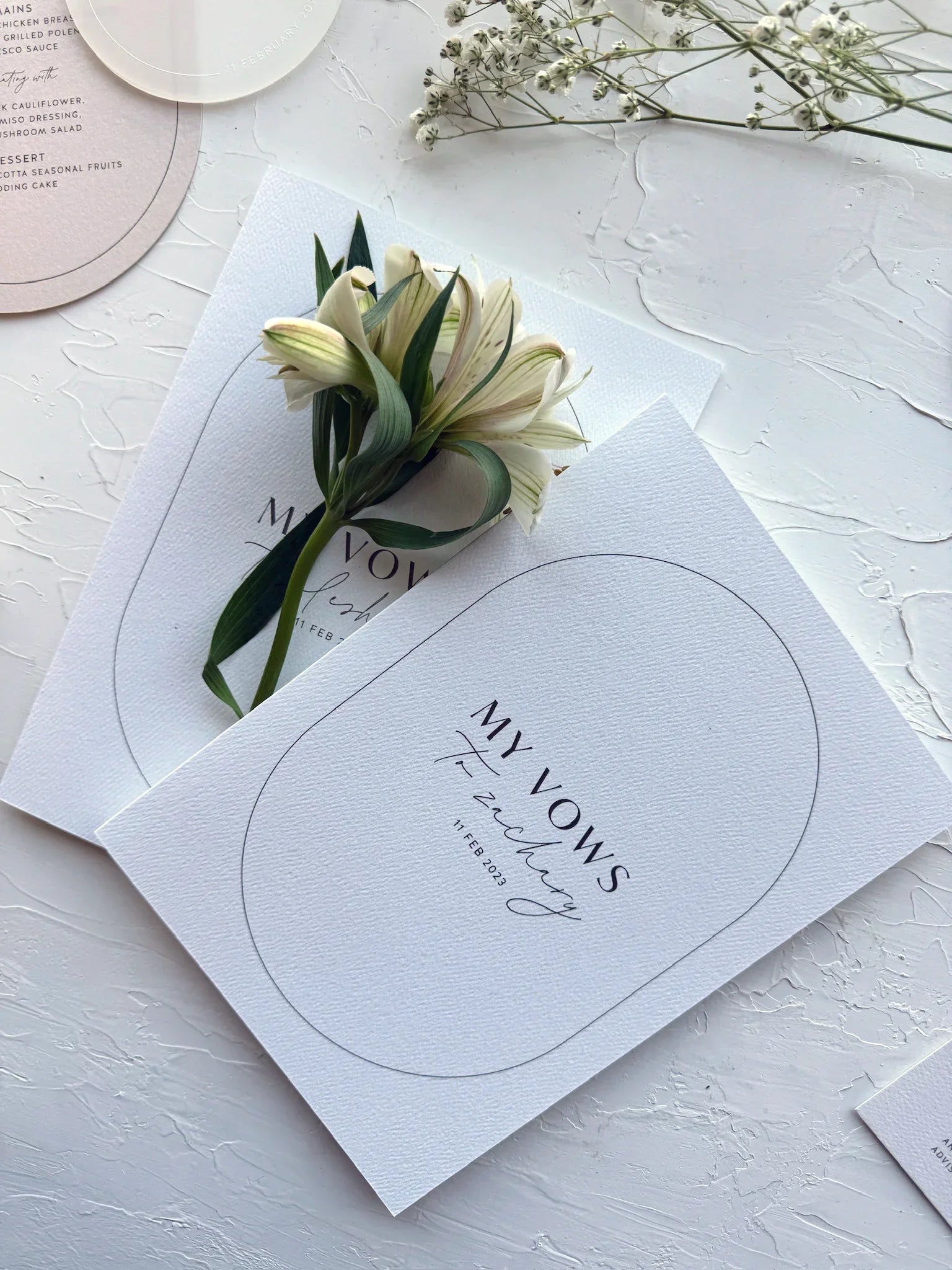 Elegant wedding vow booklets with white textured covers and delicate flower on white textured background