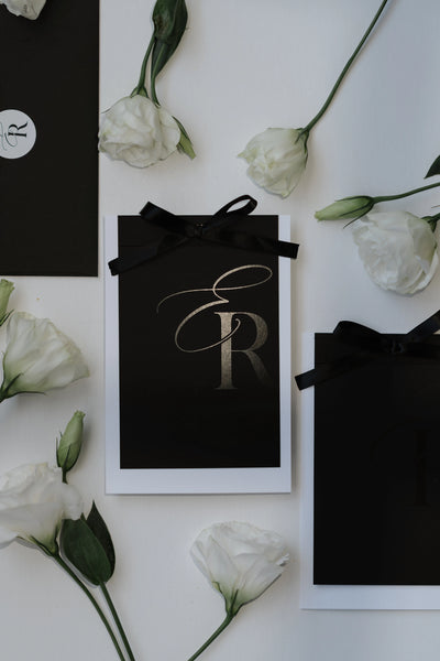 Tailored Touch black wedding invitation card with gold foil ER monogram and black ribbon bow surrounded by white lisianthus flowers by Felicitations