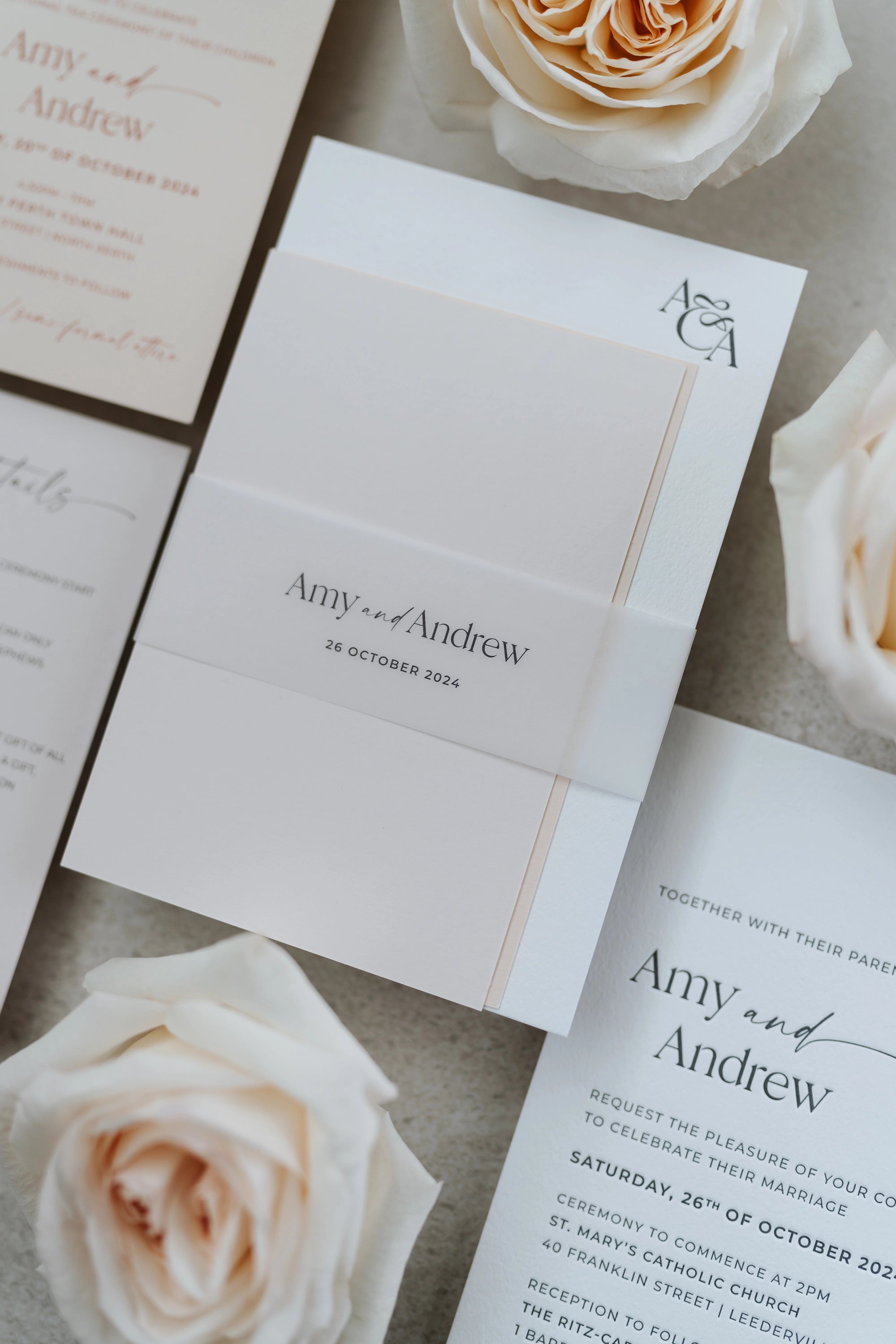Wedding invitations with 'Amy and Andrew' text, surrounded by white roses on a textured surface.