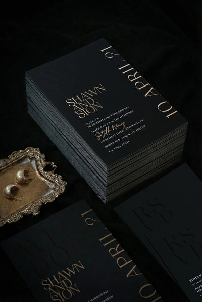 Stack of black Monaco wedding invitations with rose gold foil for Shawn and Sion, ornate vintage tray with gold half-moon earrings beside