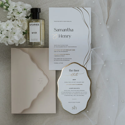 The Positano suite flat-lay with Samantha and Henry invitation, gold-edged details card, blush wave-cut envelope, perfume and white stock blooms on stone