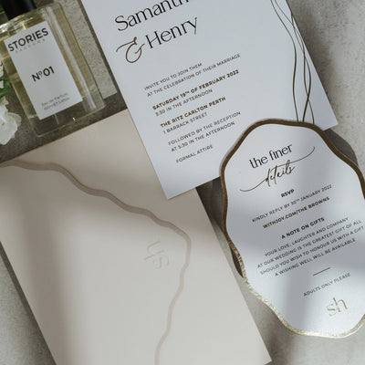 The Positano wedding suite overhead detail — gold foil wave-shape details card paired with main invitation and wave-cut blush envelope on textured stone background