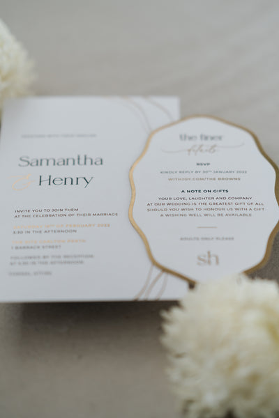 The Positano wedding invitation suite styled close-up showing gold foil typography on white invitation and ornate scallop-edge details card