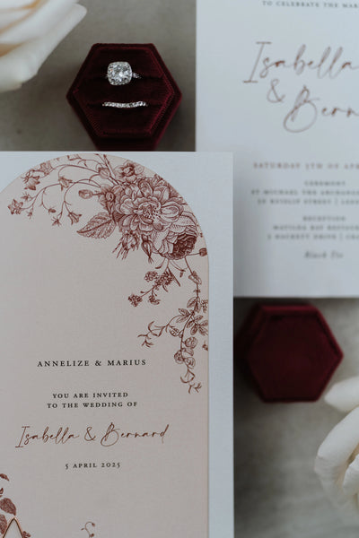 The Lisbon arch-shape invitation close-up showing detailed burgundy floral illustration on blush card with ring box and white roses styling