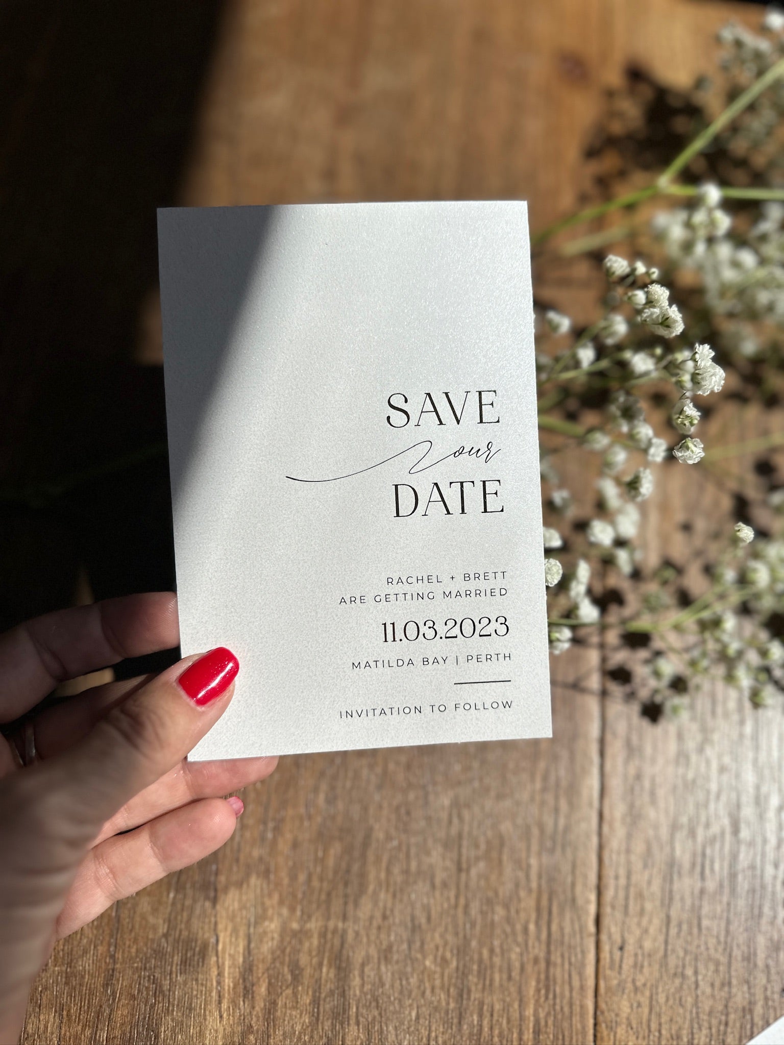 The Santorini Suite shimmer Save Our Date card for Rachel and Brett held over baby breath florals by Felicitations